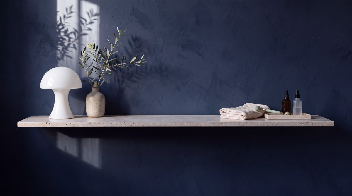 Travertin-Wandregal mit Opalglas-Lampe, Olivenzweig und Pflegeprodukten vor navyblauer Wand im Morgenlicht
