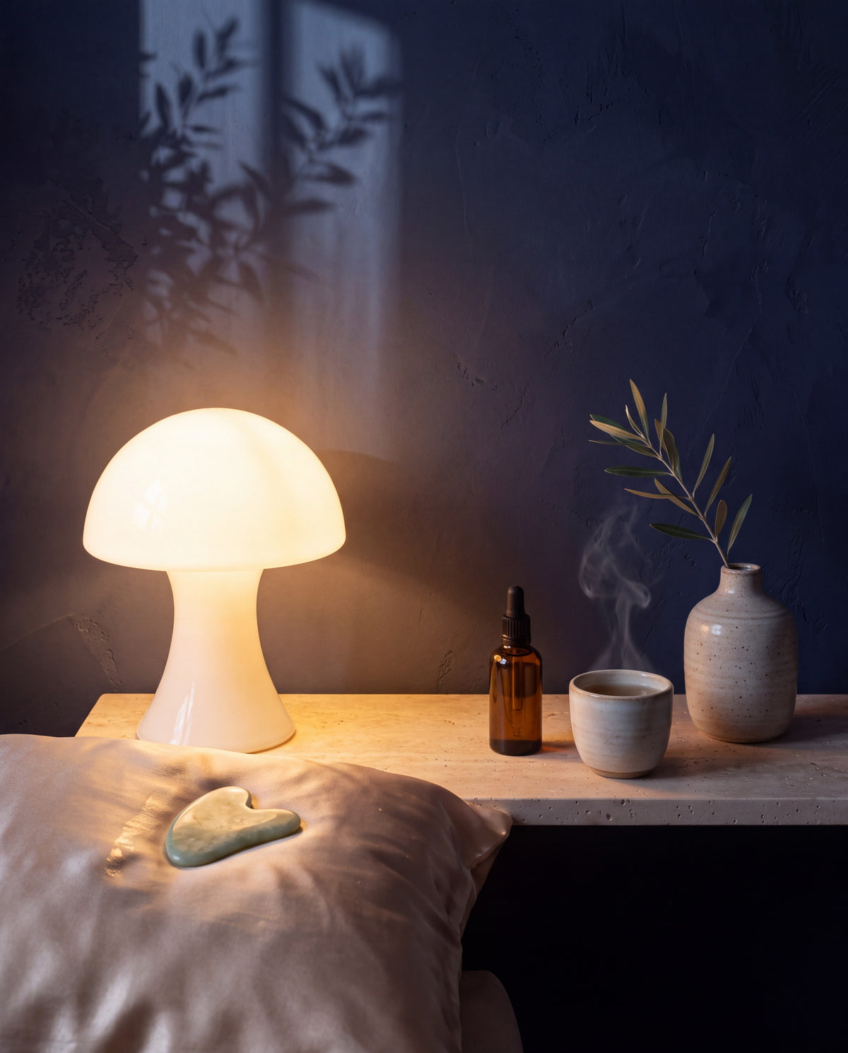 Abendritual: leuchtende Opalglas-Lampe, Seidenkissen mit Gua-Sha-Stein, Tee und Serum vor navyblauer Wand